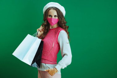 Koronavirüs salgını sırasında hayat. Pembe kolsuz gömlekli, pembe tıbbi maskeli ve yeşil arka planda izole edilmiş alışveriş çantalı modern kadın portresi..
