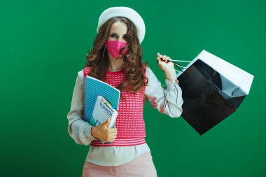 Covid-19 salgını sırasında hayat. Ders kitapları, defterleri, pembe sağlık maskesi ve yeşil arka planda alışveriş torbaları olan beyaz bereli neşeli, modern bayan öğrenci..