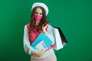 Koronavirüs salgını sırasında hayat. Beyaz bereli, ders kitapları, not defterleri, pembe tıbbi maske ve yeşil arka planda izole edilmiş alışveriş çantaları olan modaya uyan kadın portresi..