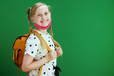 Koronavirüs salgını sırasında hayat. Beyaz puantiyeli bluz içinde, sarı sırt çantalı ve karatahta yeşil arka planda izole edilmiş pembe maskeli gülümseyen modern öğrencinin portresi..