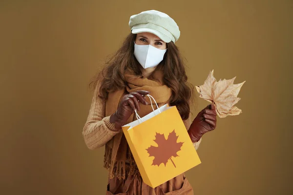 Hello autumn. smiling stylish female in sweater with white medical mask, yellow autumn maple leaves and paper shopping bag isolated on brown background.