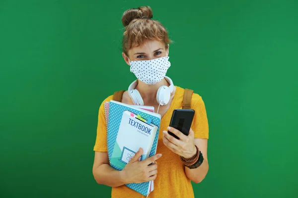 年轻的女学习者穿着黄色衬衫 带着课本 笔记本 医疗面罩 白色耳机和背包 用智能手机进行信息共享和与绿色隔离的网络联系 — 图库照片