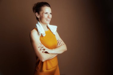 Sarı kolsuz bluzlu, kolları çapraz ve boynu havluyla sarılı, kendinden emin bir kadın, düşünceli bir şekilde sağa sola bakıyor. Rahatlamış gülüşü ve dengeli duruşu egzersizden sonra güç ve refahı yansıtıyor.