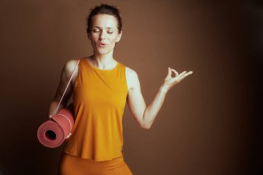 Huzurlu bir kadın, gözleri meditasyonda kapalı, pembe bir yoga minderi tutuyor ve Mudra el hareketi yapıyor. Turuncu bir bluz ve tayt giymişti. Farkındalık ve sağlığın vücut bulmuş haliydi..