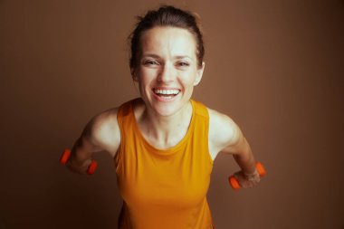 Yüksekten çekilen gülümseyen bir kadın kahverengi arka planda turuncu halterleri tutuyor. Pozitif ve enerjik tavrı spor, refah ve sağlıklı bir yaşam tarzı yayıyor..