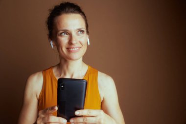 Turuncu kolsuz tişörtlü ve kulaklıklı bir kadın akıllı telefonunu tutuyor, düşünceli bir şekilde yukarı ve sağa bakıyor, teknolojiye odaklanmış bağlılığı resmediyor..