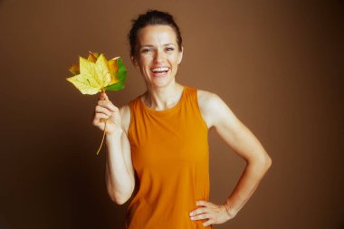 Sarı kolsuz bluzlu gülümseyen bir kadın canlı sarı ve yeşil sonbahar yaprakları tutar. Yumuşak kahverengi arkaplanla göğüs göğüse çekilen bu resim doğanın ve sonbaharın güzelliğini yansıtıyor.
