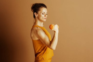 Orta yaşlı bir kadın turuncu halterle biseps kıvırıyor. Düz kahverengi arkaplana karşı yan profil çekimi fitness ve sağlıklı, aktif yaşam tarzına kararlılık ve adanmışlığı vurgular.