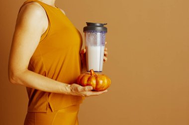 Turuncu giysili bir insanın elinde beyaz sıvı protein karıştırıcısı ve küçük bir sonbahar balkabağı tutarken görüntüsü, sağlığı ve mevsimsel rahatlığı simgeliyor..