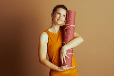 Turuncu atletik giysili gülümseyen bir kadın yoga minderini yanak yanağa yuvarlayarak kahverengi arka plana doğru bakıyor. Huzurlu ifadesi, düşünceli hareketlerle barışı ve sağlıklı yaşam tarzını temsil ediyor.