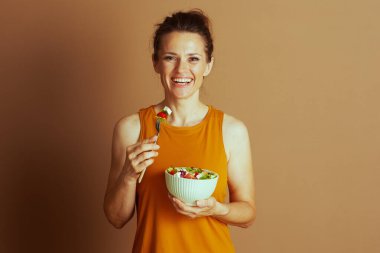 Turuncu bluzlu gülümseyen bir kadın taze salatadan domates ve feta ile çatal tutuyor. Kahverengi arka planda kameraya bakıp sağlıklı beslenmeyi teşvik ediyor..