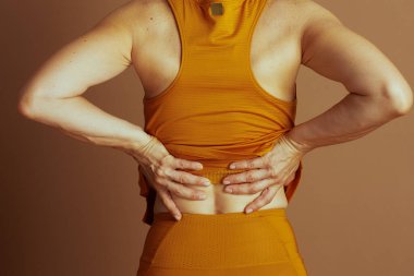 Arkadan görülen bir yetişkin, rahatsızlık ya da acı göstergesi olarak belini sıkar. Turuncu atletik bir bluz giyiyorlar ve kahverengi bir arka plana tozluk giyiyorlar. Bu da fiziksel bir gerginlik olduğunu gösteriyor..