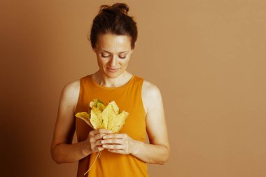 Orta yaşlı bir kadın nazik bir gülümseme ile göğsüne yakın canlı sarı sonbahar yaprakları tutar, sakin ve düşünceli bir güzelliğin vücut bulmuş halidir..