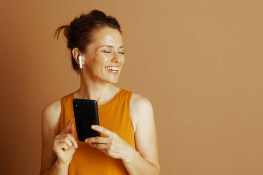 Gülümseyen orta yaşlı bir kadın, gözleri kapalı, beyaz kulaklıklı ve akıllı telefonlu kişisel seslerden hoşlanıyor. Hardal sarısı bir kolsuz bluz giyerek, bronz bir arkaplan karşısında memnuniyet yayıyor..