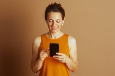 Çörekli gülümseyen bir kadın akıllı telefonuna odaklanmış turuncu ve kablosuz kulaklık takıyor. Ekrana dokunur, düz kahverengi bir arka plana karşılık gelişigüzel bir ilişki ve memnuniyet iletir..