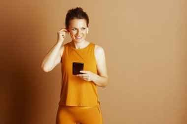 Aktif giyimli neşeli bir kadın kulaklığını ayarlıyor, elinde akıllı bir telefon var, egzersiz ya da dijital müdahale için hazır. Gülüşü aktif, sağlıklı ve bağlı bir yaşam tarzı yayıyor..