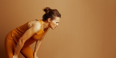 Turuncu giysili kararlı bir kadın antrenman molasında dizlerinin üstüne çöküp öne eğiliyor. Odaklanmış ifadesi, vücut geliştirmede çaba, yorgunluk ve dayanıklılık gösteriyor..