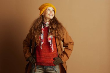 Sarı şapkalı ve turuncu kazaklı bir kadın, elleri kalçasında, yukarı bakarken kendinden emin bir şekilde gülümsüyor. Sıcak sonbahar katmanları giyinerek, tarafsız arka plana karşı açık hava faaliyetleri için iyimserliğini iletiyor.