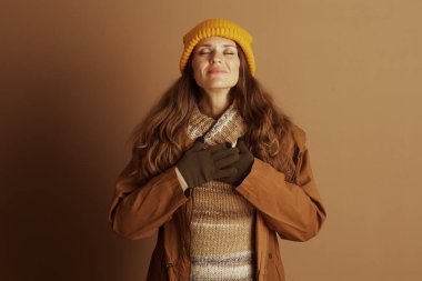 Sarı örgü şapkalı ve sonbahar katmanlı bir kadın, gözlerini huzurlu bir gülümsemeyle kapatıyor, elleri göğsüne yapışıyor, doğanın içinde huzur, minnettarlık ve iç yansıma hissi uyandırıyor..