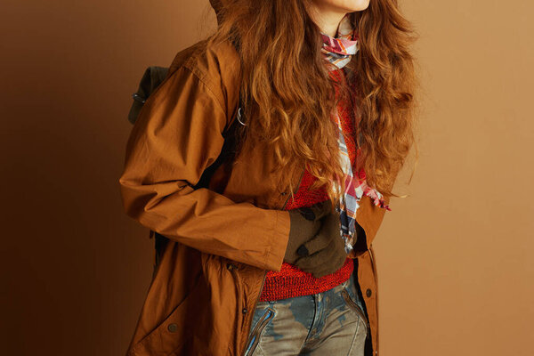 A woman in a stylish layered autumn outfit, including an orange sweater, brown jacket, and plaid scarf, stands with hands clasped, ready for cold weather or outdoor adventure.