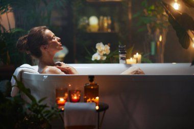 30-40 yaşlarında bir kadın beyaz köpük banyosunda huzur bulur gözleri kapalı, elleri göğsünde dinlenir. Bitkiler, mumlar ve kehribar şişeleri hoşgörü ve kişisel bakım için sakin ve rahat bir ortam oluşturur..