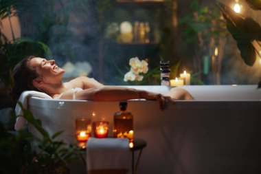 30-40 yaşlarında bir kadın köpük banyosunda, kafası havluda dinlenirken, gözleri kapalı gülümserken çok mutlu olur. Mumlar, dekoratif bir şişe, yeşillik ve çiçekler buharlı, yumuşak, sakin bir spa atmosferi yaratır..