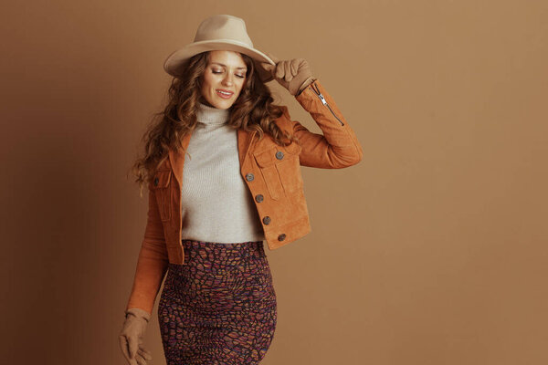 Elegant woman with curly brown hair, tan fedora, orange suede jacket, and grey turtleneck, poses confidently with hand on hat. Her stylish autumn outfit stands out against solid brown backdrop.