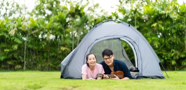 Kız arkadaşı için gitar çalan Asyalı genç bir adamın portresi. Mutlu genç çift kamp ateşinin yanında oturup gitar çalıyor.