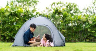 Kız arkadaşı için gitar çalan Asyalı genç bir adamın portresi. Mutlu genç çift kamp ateşinin yanında oturup gitar çalıyor.