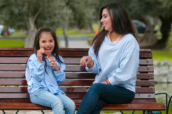 Madre y su hijita sentadas en un banco comiendo una galleta — Foto de Stock