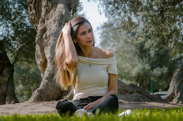 Schöne brünette Frau, die ihre langen Haare berührt. sie sitzt in einem Park auf grünem Gras. — Stockfoto