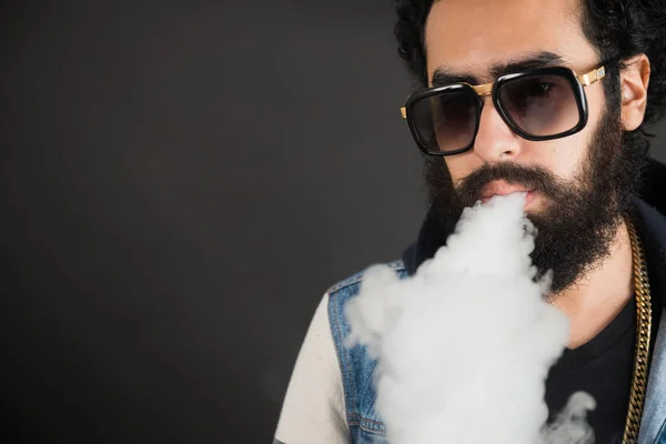 Genç vaping, stüdyo çekim adam. Bununla birlikte güneş gözlüklü bir bulut duman siyah arka plan üzerinde üfleme sakallı adam. Kavramı yasaktır ve nikotin olmadan Buhar.