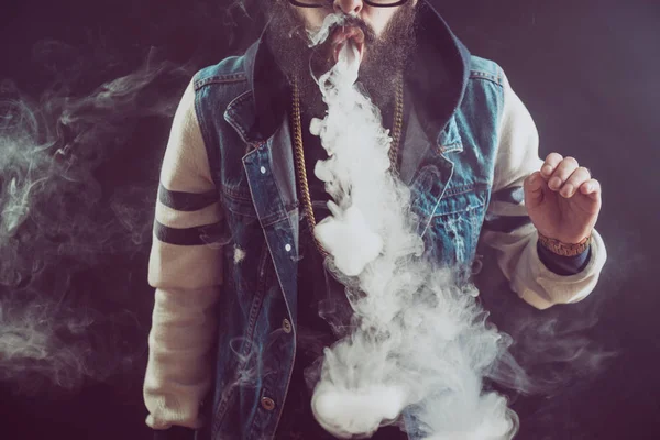 Un joven con barba arrojando una nube de vapor. Fondo negro. Vapear un cigarrillo electrónico —  Fotos de Stock