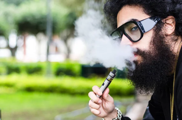 El hombre barbudo fuma vapor de cerca. Concepto de cigarrillo electrónico. Hombre con barba larga y nubes de humo se ve relajado . — Foto de Stock