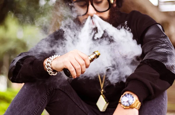 Man med skägg röker en elektronisk cigarett sitta på en bänk — Stockfoto