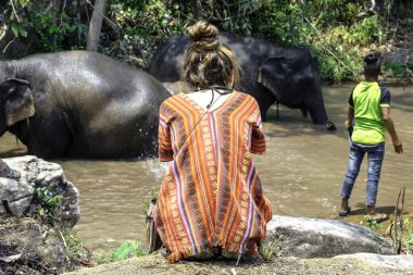 Genç bir turist sırt üstü oturmuş, köylülerle birlikte filleri seyrediyor..