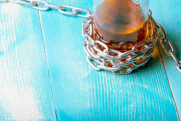 A bottle with a brown liquid around it is wound around a chain, on a wooden table