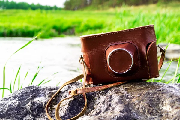 old camera in a brown case on a background of green grass and river. Travel with film camera