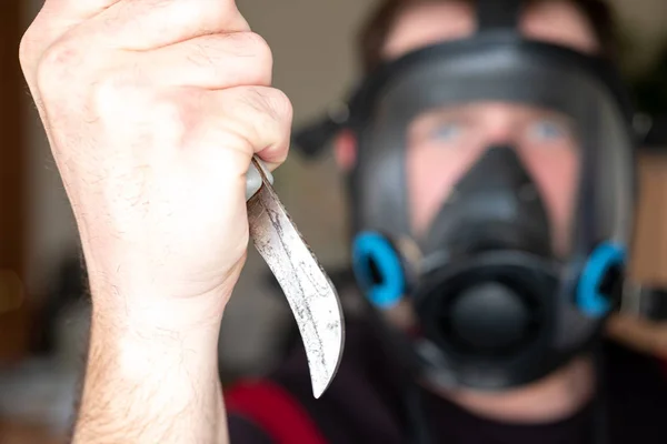 A hand with a curved knife in the foreground and a man in a gas mask in the background out of focus — Stock Photo, Image