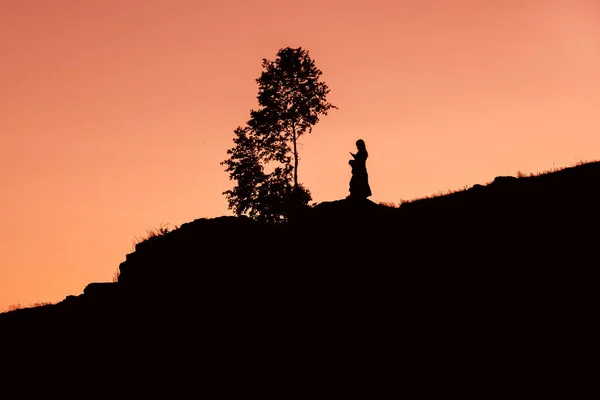 Foto av en mystisk flicka i en mörk nyckel på ett berg nära ett träd mot solnedgången himlen — Stockfoto