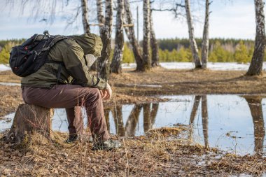 Yorgun bir adam gaz maskesi ve kapüşonlu bir kütüğün üzerinde oturuyor ve doğada solunum korumasıyla yürüyor. Radyasyon bölgesinde bahar yürüyüşü