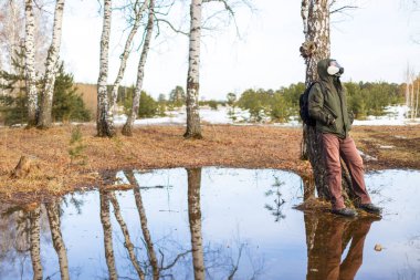 Koruyucu gaz maskeli bir adam sırtını bir huş ağacının gövdesine yaslıyor ve yukarı bakıyor. Temiz hava rüyası