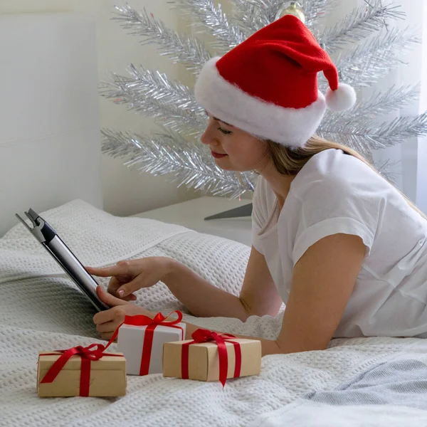 Smiling woman in santa hat is lying on the bed and using tablet for Christmas online shopping or communication