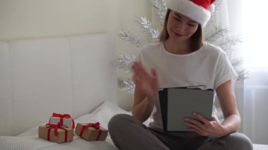 Noel çevrimiçi tebrikler. Noel Baba şapkalı neşeli kız yatakta oturup tablet kullanarak video görüşmesi yapıyor.