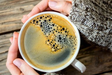 Köpük ile kokulu sıcak kahve bir fincan, eski bir dokulu ahşap masanın arka planında örme kazak kadın eller tarafından düzenlenen