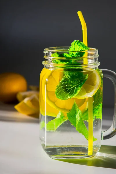 A close-up vertical high contrast photo of a mug filled with water, sliced lemon and mint leaves with yellow straw, next to a slices of lemon