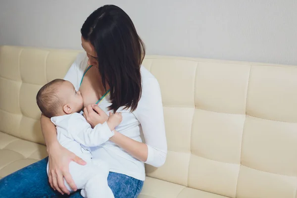 Una madre amamantando a su pequeño bebé en sus brazos. sofá beige, casa, parte superior blanca —  Fotos de Stock