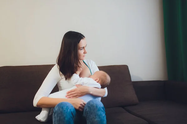 Una madre amamantando a su pequeño bebé en sus brazos. sofá marrón, casa, parte superior blanca —  Fotos de Stock