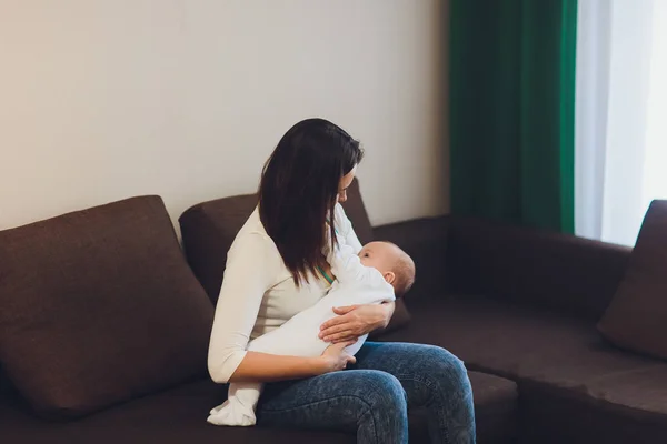 Una madre amamantando a su pequeño bebé en sus brazos. sofá marrón, casa, parte superior blanca —  Fotos de Stock