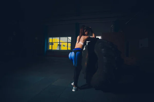 健身女子在健身房翻动车轮轮胎。适合女运动员用一个巨大的轮胎锻炼。后面的视图。做力量运动训练的运动员. — 图库照片